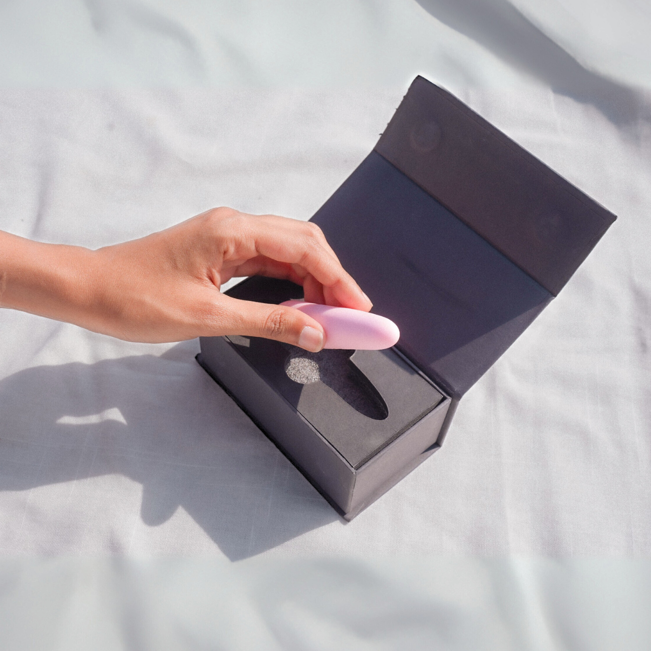 Hand holding a pink bullet massager above an open black box on a light gray background