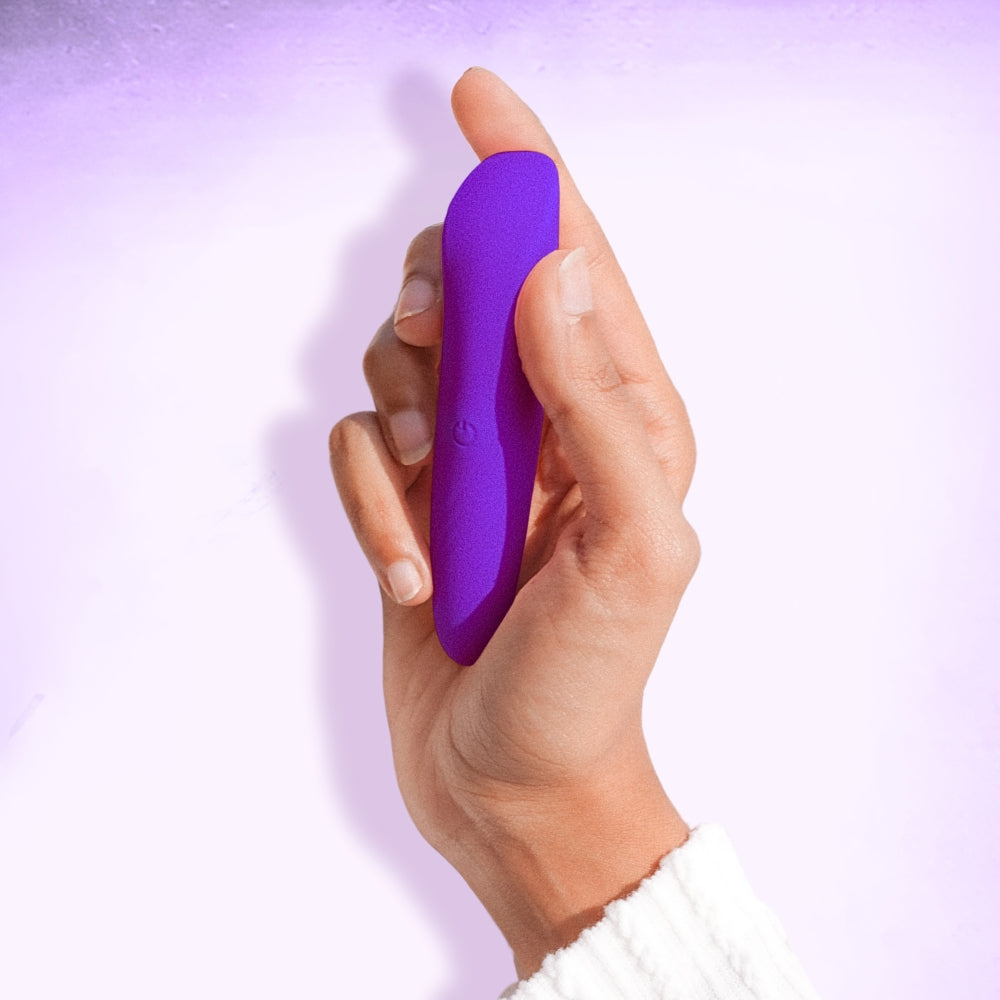 Hand holding a purple bullet massaging device against a light purple background, showcasing the product's vibrant colors