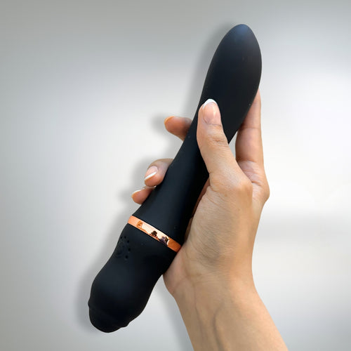 Black handheld massager with rose gold accents held in a hand against a light gray background. Sleek, smooth texture of the silicone.